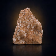 A raw mineral specimen or crystal cluster with prominent clear and pale pinkish-white crystalline structures on a warm tan-brown matrix, photographed against a dark black background with a subtle warm glow behind it.
